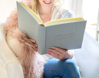May include: A gray linen-covered photo album with a yellow spine and gold lettering. The album is open and the text on the cover reads "JAMES & AYSHA OUR ANNIVERSARY 11 DECEMBER 2023".