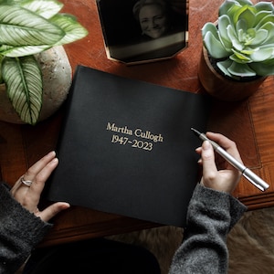 May include: A black memorial book with gold lettering that reads "Martha Culloch 1947-2023". A person is holding a pen over the book. The book is on a wooden table with a framed photo and potted plants.