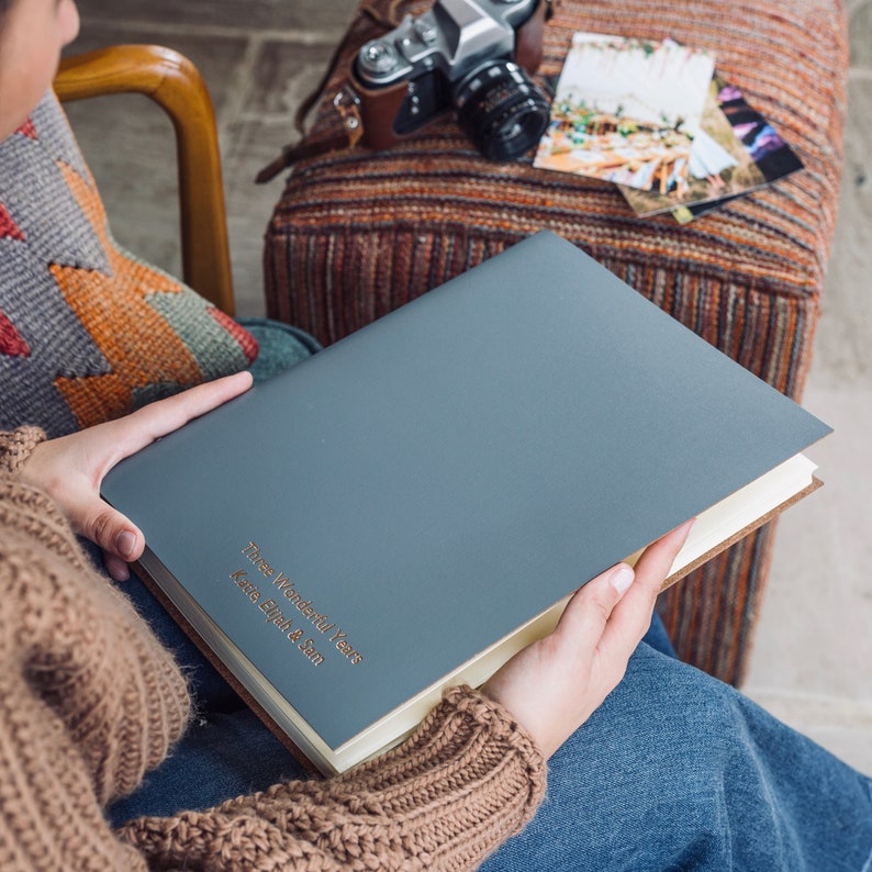 a woman is holding an A4 sized grey leather album which has been personalised with names on thr front. There is a camera on the side and some wedding photos. She is wearing jeans and a jumper