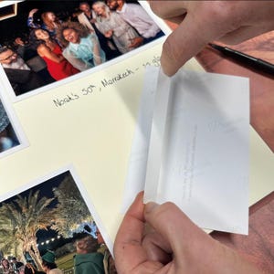 A piece of double-sided mounting tape is being applied to the back of a photo before being stuck into an open photo album.