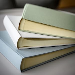 A pile of three linen photo albums are stacked up on a table. You can see the spines and all the pages inside. The albums are grey, green and white. The setting is quite stark.