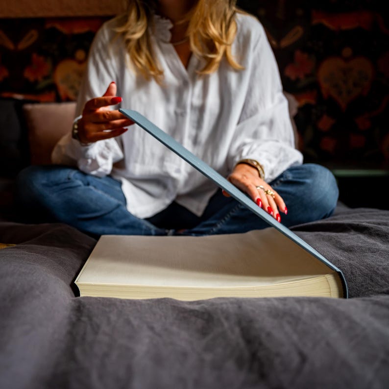 A young woman is sitting on a bed and she has a big linen photo album in her hands. The bed is dark grey and the woman is wearing a white shirt.