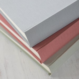A close up view of three linen photo albums where you can see the spines of the books and the pages inside. The one on the bottom is green, then a muted pink, then a pale grey.