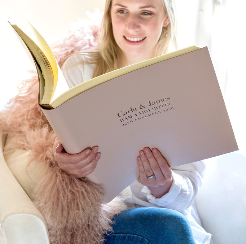 a young woman holds a large leather photo album which has been personalised with wedding details on the front. The leather photo album is a pale blush pink and the embossing is done in gold foil
