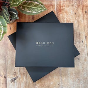 A pile of navy blue presentation boxes for photo albums or guest books sits on a wooden table top, next to a plant. The boxes are embossed with the name of the bespoke stationery company, BeGolden.