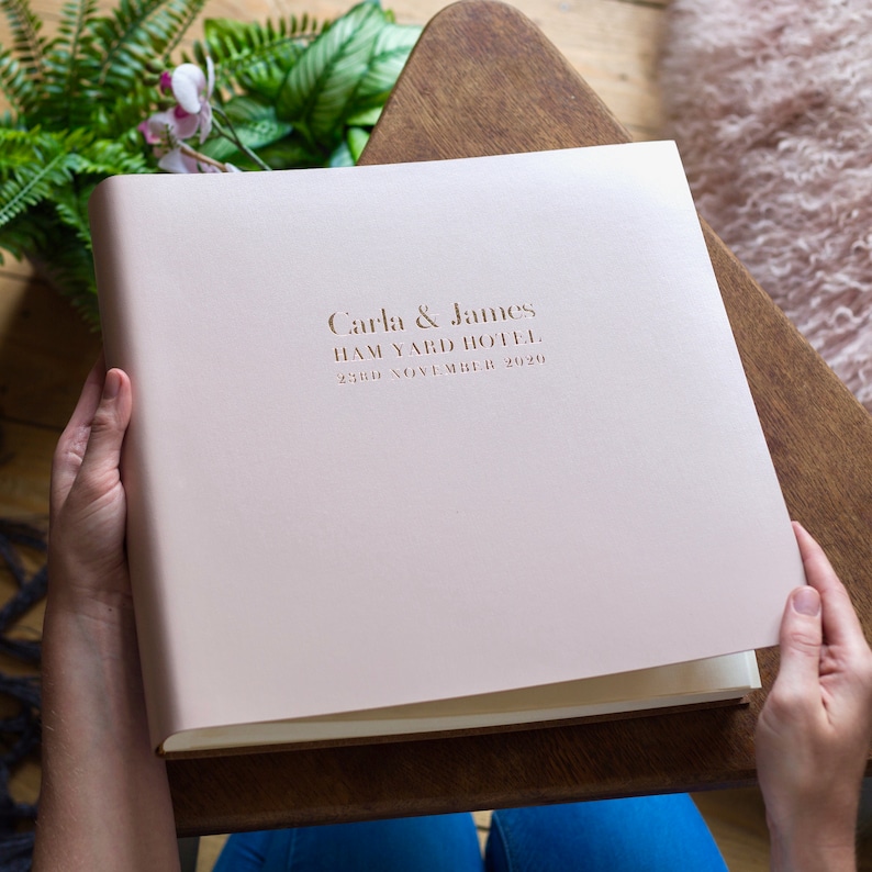 the blush pink large wedding photo album is on a wooden coffee table surrounded by a plant and a rug. The writing on the front is for a couple who have recently been married and hotfoil printed in gold foil