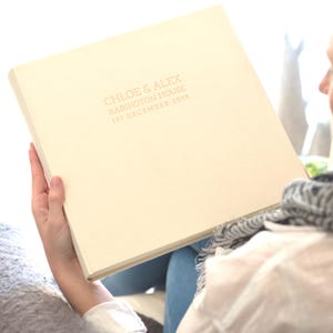 a large linen wedding photo album is being held by a person sitting in a chair. The photo album has been personalised on the front with rose gold writing.
