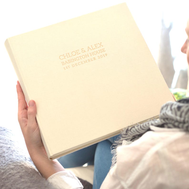 a large linen wedding photo album is being held by a person sitting in a chair. The photo album has been personalised on the front with rose gold writing.