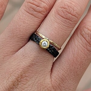 May include: A black and gold ring with a small, clear gemstone. The ring is worn on a person's finger.