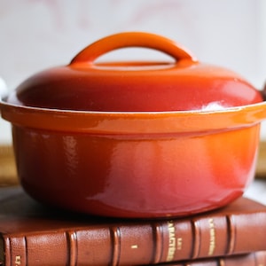 Peut inclure: Cocotte en fonte émaillée orange avec un couvercle et deux poignées. La casserole est posée sur deux livres reliés en cuir.