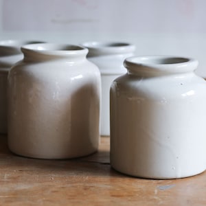 Puede incluir: Cuatro tarros de cerámica blanca con un acabado liso y mate. Los tarros están dispuestos en una fila sobre una superficie de madera.