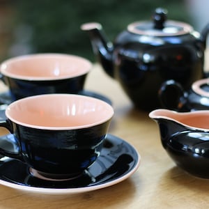 May include: A black tea set with a teapot, creamer, sugar bowl, and two teacups and saucers. The cups and saucers have a pink interior.