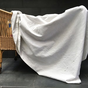 Könnte beinhalten: Ein Korbwiege mit einem weißen Laken darüber. Die Wiege hat ein geflochtenes Design und steht auf Rädern. Das Laken hat ein dekoratives Design in der Mitte.