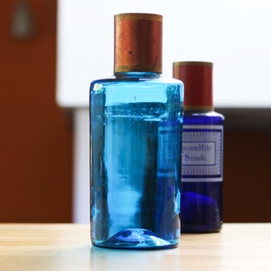 Può includere: Due bottiglie di vetro con liquido blu e tappi in rame e rosso. La bottiglia a sinistra ha un'etichetta blu con il testo "Eau de Cologne" e "Sombre".