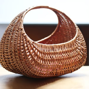 May include: A woven wicker basket with a large, oval-shaped body and a circular handle. The basket is light brown and has a natural, rustic look.
