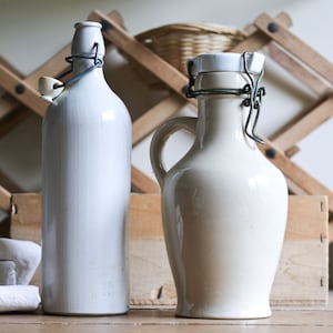 May include: Two ceramic bottles with metal closures. One is tall and slender, the other is a jug shape with a handle. Both are off-white. A wooden wine rack and a basket are in the background, suggesting a rustic or vintage theme.