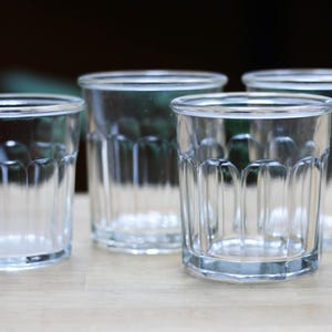 Puede incluir: Cuatro vasos de vidrio transparente para beber con un diseño acanalado. Los vasos están dispuestos en una fila sobre una superficie de madera.
