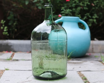 Puede incluir: Una botella grande de vidrio transparente con un tinte verde, llena de agua, se encuentra sobre una superficie de piedra. Una jarra de cerámica azul claro está en el fondo. La botella tiene forma redondeada y cuello estrecho.
