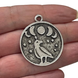 May include: Silver charm featuring a crow and moon phases design. The crow is facing right and the moon phases are in a crescent shape. The charm is round and has a textured surface.