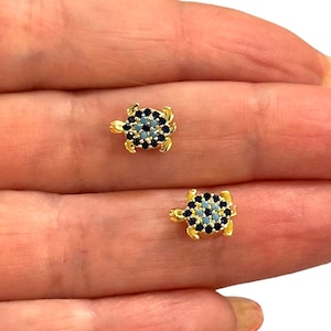 May include: A pair of gold-coloured turtle stud earrings. Each turtle features a light blue shell with a pattern of dark blue stones. The earrings are small and delicate, suitable for everyday wear.