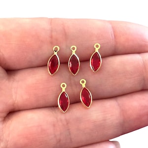 May include: Six gold-colored marquise-shaped charms with a vibrant red gemstone center. Each charm has a small loop at the top for attaching to jewelry. The charms are displayed against a light background.