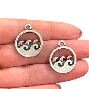 May include: Two silver-toned metal charms in the shape of a circle with a wave design inside. The charms have a loop at the top for attaching to a necklace or bracelet.
