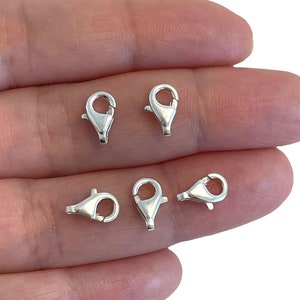 May include: Six silver lobster claw clasps, a type of jewellery finding, are shown on a hand. The clasps are used for attaching jewellery chains or cords.