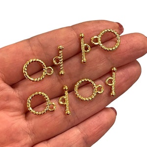 以下が含まれることがあります： 金色のトグルクラスプがいくつかあります。クラスプは、ねじれたロープのデザインの円形リングと、小さなループが付いたバーを備えています。これらのクラスプは、ジュエリー作りに使用されます。