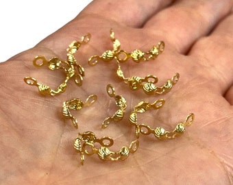 May include: A close-up of a hand holding a group of gold-colored metal clasps. The clasps are shaped like a scallop shell and have two small loops on each side.