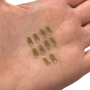 May include: A selection of small, gold-coloured metal clasps, each with a rounded end and a hole for attachment. The clasps are arranged on a light-toned background, highlighting their shiny, metallic finish. These clasps are likely used for jewellery making or crafting.