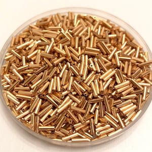 Könnte beinhalten: Nahaufnahme eines durchsichtigen Plastikbehälters gefüllt mit goldfarbenen Glasperlen. Die Perlen sind klein und zylindrisch geformt.