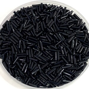 May include: A close-up view of a bowl filled with black glass seed beads. The beads are small and cylindrical in shape.