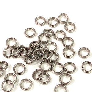 Puede incluir: Un conjunto de pequeños anillos divididos de color plateado dispersos sobre una superficie blanca. Estos anillos metálicos son circulares con una división, lo que permite abrirlos y sujetarlos a otros objetos. Los anillos se utilizan probablemente para la fabricación de joyas o manualidades.