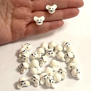 May include: A group of white ceramic mouse head buttons with black eyes and pink cheeks. The buttons have a hole in the back for sewing.