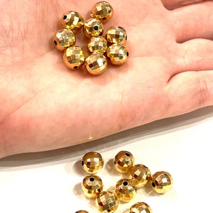 May include: A close-up of a pile of gold-colored, faceted round beads. The beads are scattered on a hand, with more beads scattered on a white surface below.