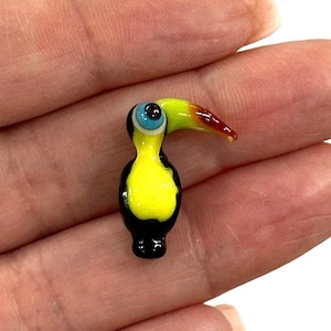 Puede incluir: Una pequeña figura de tucán de vidrio con cuerpo amarillo, cabeza y alas negras, pico rojo y amarillo y un ojo azul.