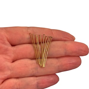 Puede incluir: Un conjunto de alfileres dorados con pequeñas cabezas planas. Los alfileres son finos y rectos, dispuestos en un haz suelto. Los alfileres se sostienen en la mano de una persona sobre un fondo blanco.