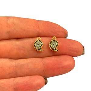 May include: Two gold-colored charm pendants in a paisley shape. Each charm features a central turquoise-colored circle surrounded by a ring of black stones. The pendants are held in a person's hand against a white background.
