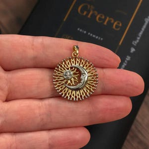 Può includere: Un ciondolo dorato con un design a sole e luna. Il sole è al centro, con una faccia sorridente sulla luna. Il ciondolo ha una cornice circolare dettagliata con un anello per una catena.