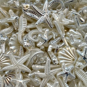 Puede incluir: Primer plano de una colección de adornos de conchas marinas y estrellas de mar nacaradas y plateadas. El surtido incluye varias formas y tamaños de estrellas de mar, conchas marinas e hipocampos, creando una apariencia texturizada y detallada.
