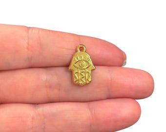 May include: A gold-colored Hamsa hand charm with an eye design. The charm is small, with intricate details, and has a loop at the top for attaching to jewelry. The charm is held in a person's hand.