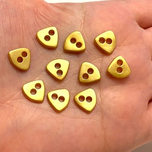 May include: Twelve gold triangle-shaped buttons with two holes each. The buttons are arranged in a semi-circle on a hand.