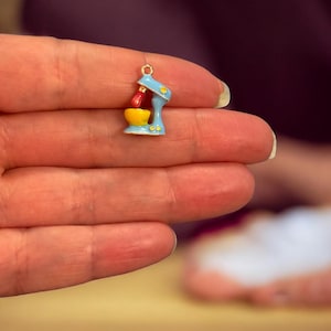 May include: A small, brightly coloured charm depicting a stand mixer. The mixer is painted light blue, with a yellow bowl and a red beater. It has a gold-coloured loop at the top, suitable for jewellery making.