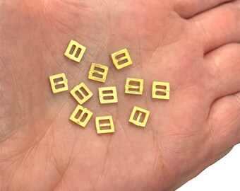 May include: A close-up of a hand holding a group of small, gold-colored metal buckles. The buckles are square-shaped with two holes on each side.