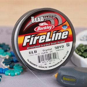 May include: A spool of FireLine microfused braided bead thread by The Beadsmith. The label shows the brand name, thread type, and weight of 6LB. The spool is gray with a red, yellow, and white label. A glue gun and beads are in the background.