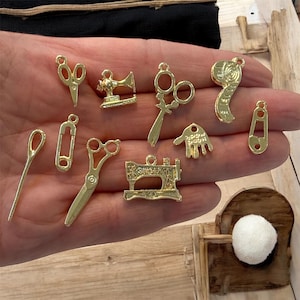 May include: A selection of gold-coloured sewing-themed charms, including scissors, sewing machines, safety pins, and a spool of thread. These small, detailed charms are ideal for jewellery making or craft projects. The charms are displayed against a light wooden surface.