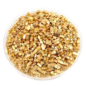 May include: A close-up view of a bowl filled with gold-colored metal beads. The beads are small and rectangular in shape, with two holes on each side. The beads are arranged in a random pattern, creating a visually appealing texture.