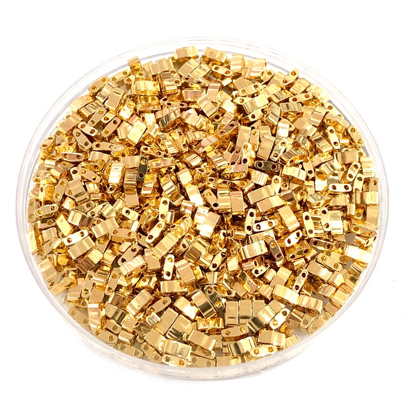 May include: A close-up view of a bowl filled with gold-colored metal beads. The beads are small and rectangular in shape, with two holes on each side. The beads are arranged in a random pattern, creating a visually appealing texture.