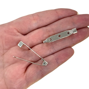May include: Silver tone pin back and safety pin. The pin back is a long, rectangular piece of metal with three holes. The safety pin is a traditional style with a pointed end and a spring.