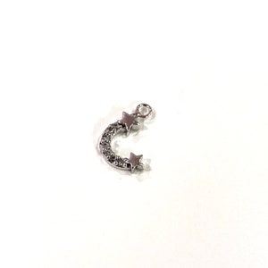 May include: Silver charm with a crescent moon shape and two star accents. The moon is decorated with small, clear crystals.
